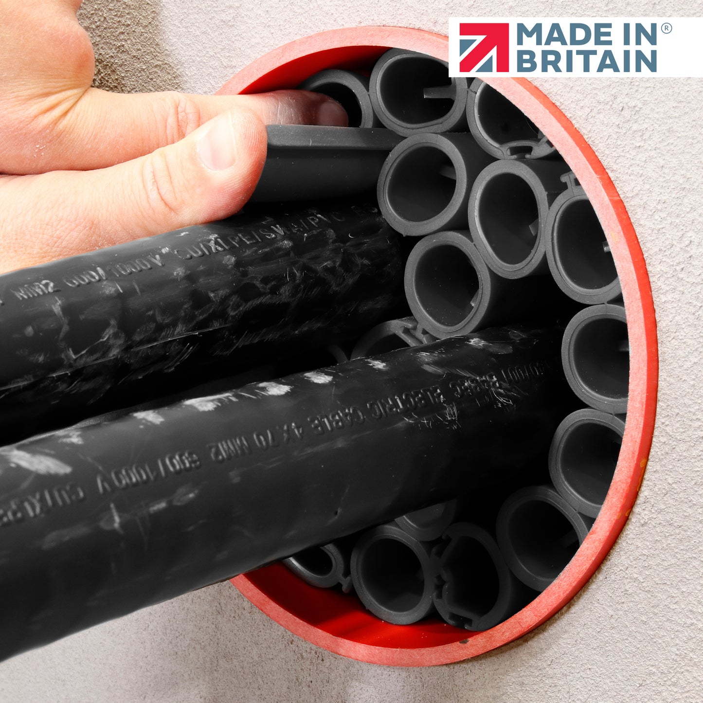Hands positioning black cable ducts inside a circular wall opening lined with a red frame, preparing for fire-rated duct sealing. Made in Britain logo displayed.