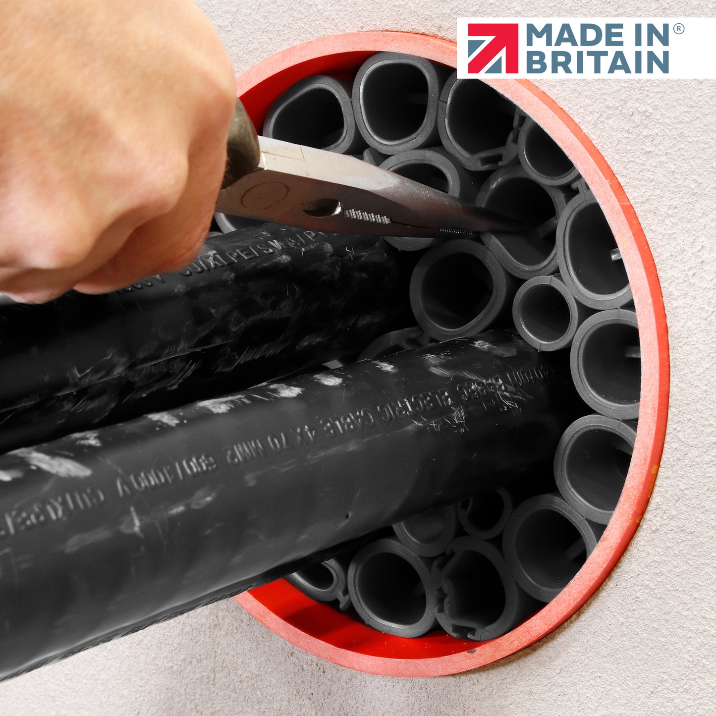 Installer cutting black cable ducts with scissors inside a red circular wall sleeve during fire-rated sealing preparation. Made in Britain logo visible.