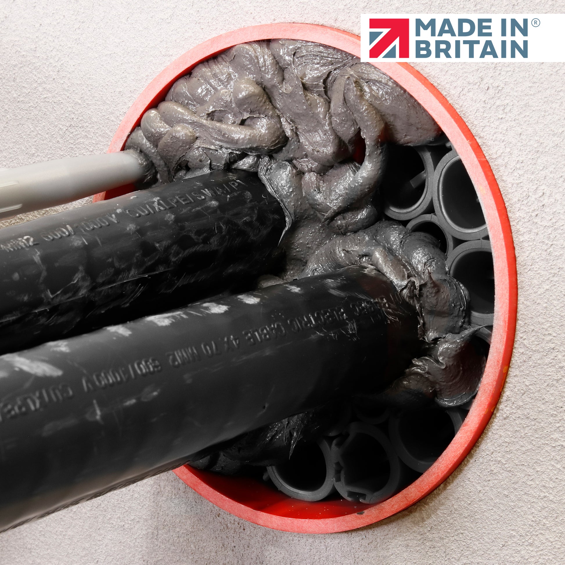 Close-up of DuctSeal FR fire-rated sealant applied around black cable ducts within a red-sealed circular wall opening, showing dense grey fireproof sealant and the ‘Made in Britain’ logo.
